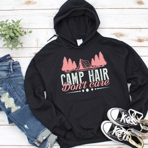K&ouml;nnte beinhalten: Schwarzer Kapuzenpullover mit dem Text "CAMP HAIR Don't care" in Wei&szlig; und Rosa. Das Design zeigt ein rosa Zelt und B&auml;ume. Dazu blaue Jeans und schwarze Leinenschuhe.
