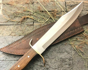 May include: A brown wooden handled knife with a silver blade and a brown leather sheath. The knife is on a stone surface with dried grass.