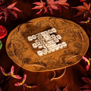 Può includere: Un tradizionale set di giochi cinese con tessere bianche con caratteri neri, disposte su un tappetino di pelle marrone con motivi di draghi. Sono visibili pezzi decorativi rossi e verdi, insieme a foglie autunnali rosse.