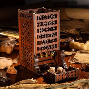 Könnte beinhalten: Ein Würfelturm aus Holz mit aufwendigen Ausschnitten und der lateinischen Inschrift "Vivas Pictos Victos Hostis Deleta Lvdite Secvri". Der Turm hat eine kleine Schale zum Auffangen der Würfel und wird von zwei Würfeln und kleinen goldenen Glocken begleitet.