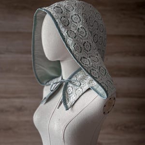May include: A light blue lace bonnet with a matching collar and ties, displayed on a mannequin. The bonnet features a floral lace pattern and is trimmed with a darker blue satin ribbon. The background is a light brown wooden wall.