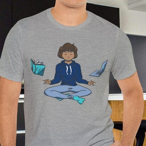 Könnte beinhalten: Ein hellgraues T-Shirt mit einem Grafikdruck einer Person, die meditiert, mit einem Laptop, einem Buch und einem Telefon, die um sie herum schweben. Der Text auf dem Shirt lautet "Zen und die Kunst des Multitaskings".