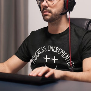 Pode incluir: Uma pessoa vestindo uma camiseta preta com texto branco que diz "Progress Incremented" e um rosto sorridente com dois sinais de mais como olhos. A pessoa está usando um fone de ouvido e olhando para uma tela de computador.