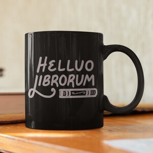 Può includere: Tazza da caffè in ceramica nera con un design bianco che dice "Helluo Librorum" con un'illustrazione di libro stilizzata.