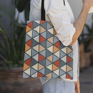 May include: A person wearing a white shirt and blue jeans is carrying a tote bag with a geometric pattern of triangles in shades of blue, orange, and beige. The bag has black straps. The person is holding a white coffee cup in their right hand.