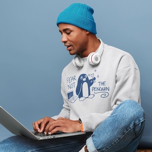 Może przedstawiać: Osoba w jasnoszarej bluzie z niebieską grafiką pingwina i napisem "Fear Not The Penguin". Osoba ma na sobie niebieską czapkę, niebieskie dżinsy i białe słuchawki. Siedzi i używa laptopa.