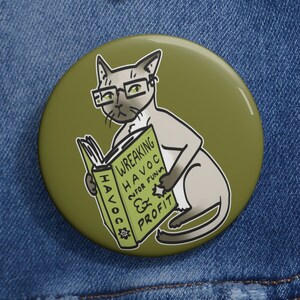 May include: A green button with a cartoon illustration of a cat wearing glasses and reading a book titled "Wreaking Havoc for Fun & Profit".