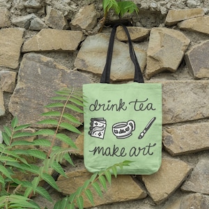 May include: A green tote bag with black handles and the text "drink tea make art" in black script. The bag has three small black and white illustrations of a book, a teacup, and a paintbrush.