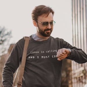 以下が含まれることがあります： 白い文字で「Linux is calling AND I MUST CODE」と書かれた濃いグレーのトレーナーを着た男性。青いジーンズとサングラスをかけ、茶色の革製のダッフルバッグを持っている。