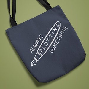May include: A navy blue tote bag with black straps and a white design of a pen with the text "ALWAYS PLOTTING SOMETHING".