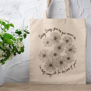 May include: A beige canvas tote bag with a black and white daisy design and the text "Daisy Daisy, give me your answer do; I'm half crazy all for the love of you."