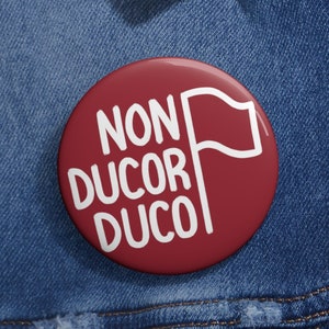 May include: A red button with white text that reads "NON DUCOR DUCO" and a white flag. The button is pinned to a blue denim jacket.