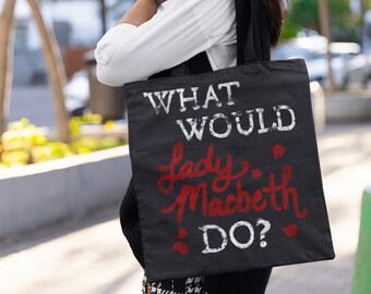 Könnte beinhalten: Eine schwarze Tragetasche mit weißem Text, der "What Would Lady Macbeth Do?" lautet. Der Text ist in einer verwitterten Schriftart gehalten. Die Tasche wird von einer Person getragen, die ein weißes Hemd, eine schwarze Weste und einen schwarz-weiß karierten Rock trägt.