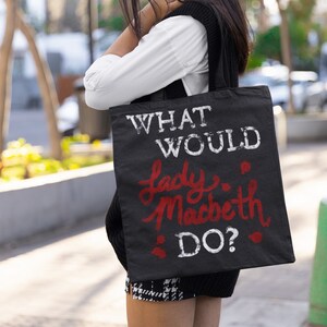 Puede incluir: Una bolsa de tela negra con texto blanco que dice "What Would Lady Macbeth Do?" El texto está en una fuente desgastada. La bolsa la lleva una persona que lleva una camisa blanca, un chaleco negro y una falda de cuadros blanco y negro.