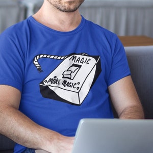 Pode incluir: Um homem vestindo uma camiseta azul com um gráfico branco de um interruptor de luz que diz "Magic" e "More Magic". Ele está sentado em um sofá cinza e usando um laptop.