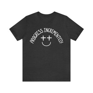 May include: A dark heather grey t-shirt with the text "Progress incremented!" in white, featuring a smiley face with two plus signs for eyes.