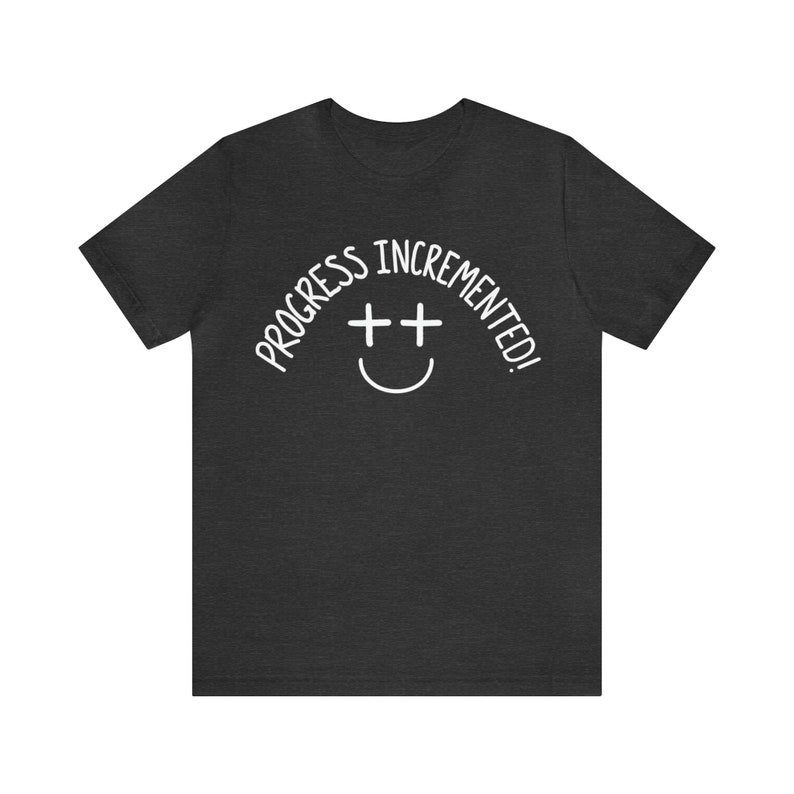 May include: A dark heather grey t-shirt with the text "Progress incremented!" in white, featuring a smiley face with two plus signs for eyes.