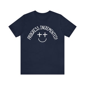 May include: Navy blue t-shirt with white text that reads "Progress incremented!" and a smiley face with two plus signs for eyes.