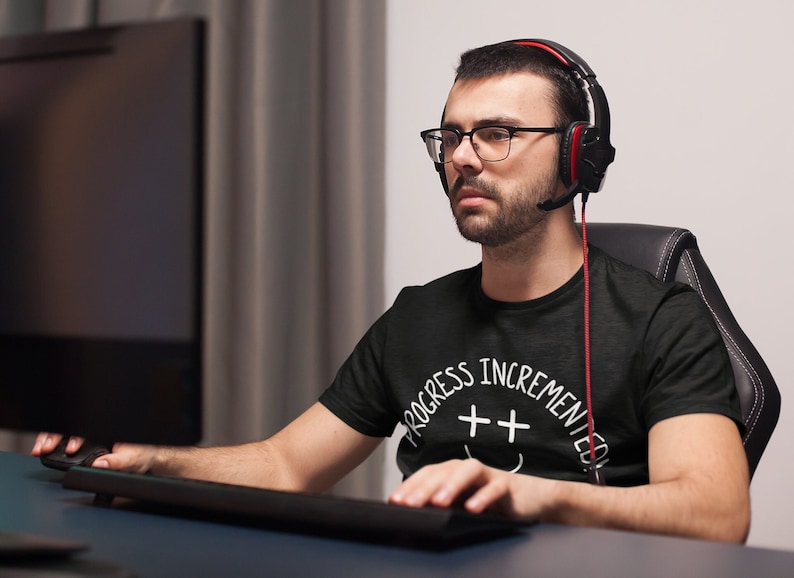 May include: A person wearing a black t-shirt with white text that reads "Progress Incremented" and a smiley face with two plus signs for eyes. The person is wearing a headset and looking at a computer screen.