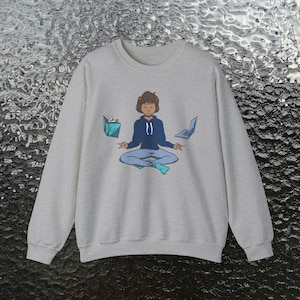 May include: A gray sweatshirt with a cartoon illustration of a person meditating with a book, laptop, and headphones. The person is wearing a blue hoodie and blue pants.