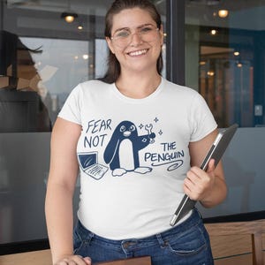 May include: White t-shirt with a navy blue graphic of a penguin holding a wrench and the text "FEAR NOT THE PENGUIN". The shirt is worn with blue jeans. The person is holding a black tablet.