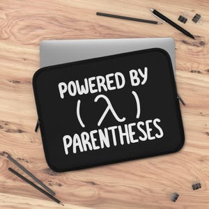 May include: Black laptop sleeve with white text reading "POWERED BY (λ) PARENTHESES". The rectangular sleeve has a zip closure. A silver laptop is partially visible behind it. Pencils and paper clips are scattered around.