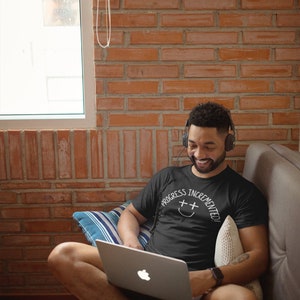 May include: A person wearing a black t-shirt with the text "Progress Incremented!" in white lettering and a smiley face with plus signs for eyes. The person is sitting on a couch and using a laptop computer.
