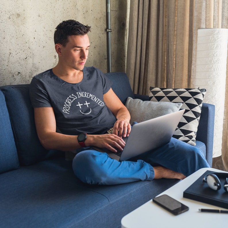 May include: A person wearing a gray t-shirt with the text "Progress Incremented" and a smiley face graphic is sitting on a blue couch and using a laptop.