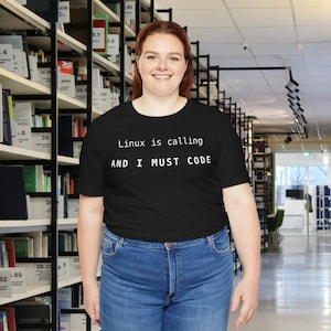 May include: Black t-shirt with the text "Linux is calling AND I MUST CODE" in white. The shirt is paired with blue jeans. The background features a library setting with bookshelves filled with books.