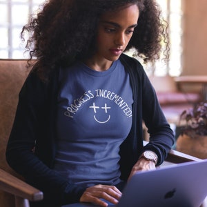 May include: A woman wearing a navy blue t-shirt with the text "Progress Incremented!" and a white smiley face with plus signs for eyes. She is sitting in a chair and using a laptop computer.