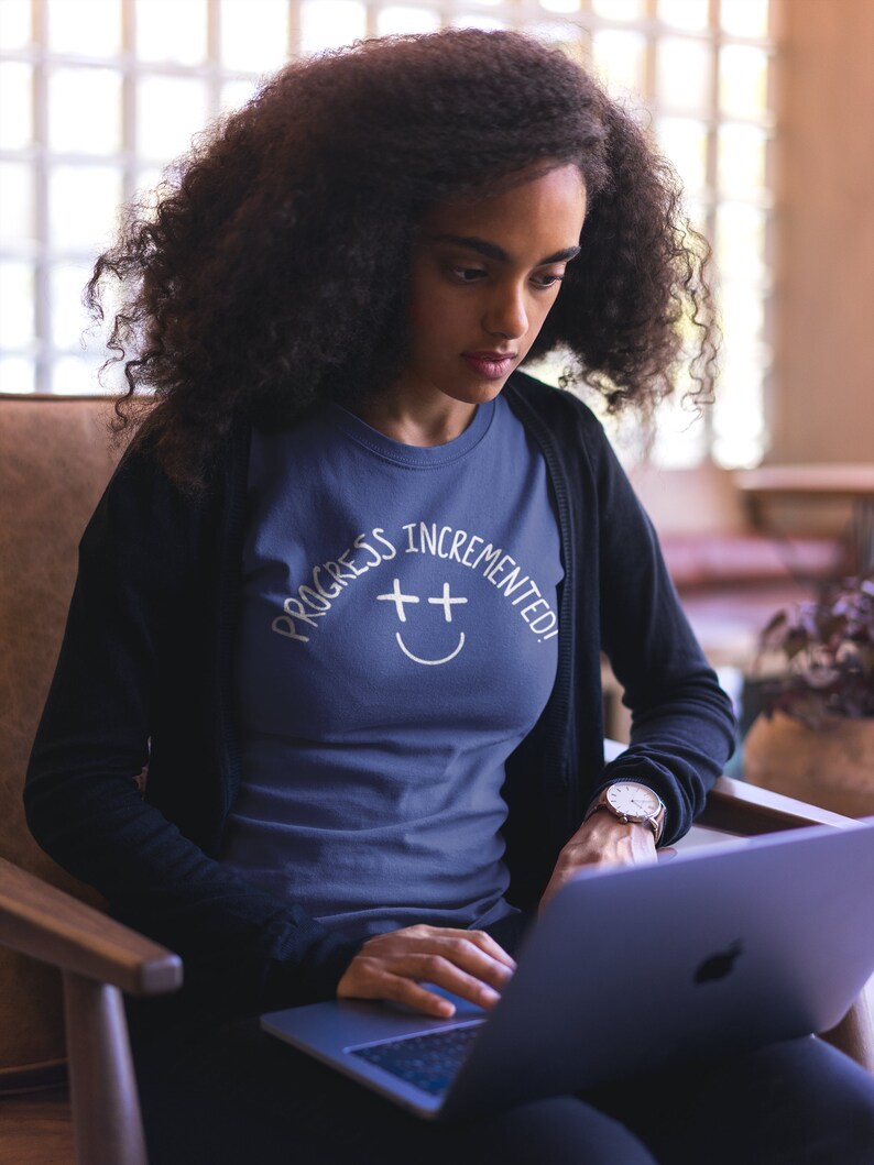 May include: A woman wearing a navy blue t-shirt with the text "Progress Incremented!" and a white smiley face with plus signs for eyes. She is sitting in a chair and using a laptop computer.
