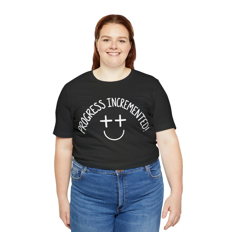 May include: A black t-shirt with white text that reads "Progress incremented!" and a smiley face with two plus signs for eyes.