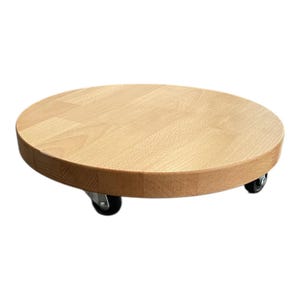 Könnte beinhalten: Ein runder Pflanzenroller aus hellem Holz mit vier kleinen schwarzen Rädern. Der Roller besteht aus hellem Holz und ist für einen Blumentopf konzipiert. Der Durchmesser des Rollers beträgt etwa 30 cm.