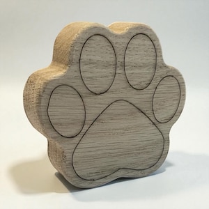 Puede incluir: Una caja de madera con forma de huella de pata, de color marrón claro con contornos grabados más oscuros. La caja tiene un diseño tridimensional, con las almohadillas y los dedos de los pies claramente definidos. Es probable que sea una pieza decorativa o un pequeño contenedor de almacenamiento.