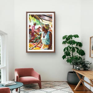 May include: Watercolor painting of a market scene. A woman in a light blue top and headscarf is arranging produce. Other figures are visible in the background. The artwork is framed in a dark wood frame and hangs on a white wall.