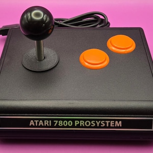 May include: Black Atari 7800 ProSystem joystick with a black ball-top handle, two orange buttons, and a silver base. The text "ATARI 7800 PROSYSTEM" is displayed on the front of the controller. A black cable extends from the back.