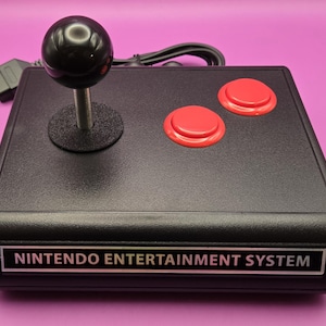 May include: A black Nintendo Entertainment System controller with a joystick and two red buttons. The controller is connected to a black cable.