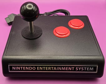 May include: A black Nintendo Entertainment System controller with a joystick and two red buttons. The controller is connected to a black cable.