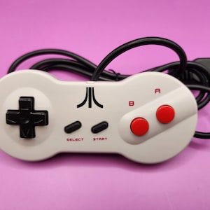 May include: A retro-style video game controller in white with a black directional pad, black buttons, and red action buttons. The controller has the Atari logo and a black cable. The background is a solid pink color.