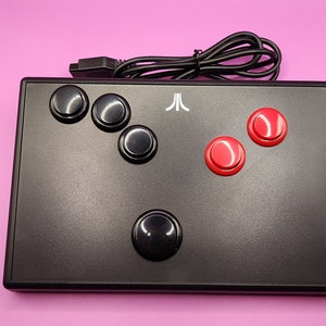 May include: Black Atari-branded arcade controller with black and red buttons. The controller has a rectangular shape with a black cable attached. The Atari logo is visible on the top of the controller.
