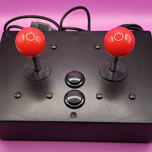 May include: Black arcade game controller with two red ball-top joysticks and two black buttons. The controller has a rectangular shape and is secured with screws. A black cable extends from the back.