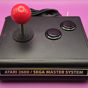 May include: Black Atari 2600 / Sega Master System joystick with a red ball-top, two black buttons, and a silver switch. The joystick has a black base with the text "ATARI 2600 / SEGA MASTER SYSTEM" in a gold-colored font.