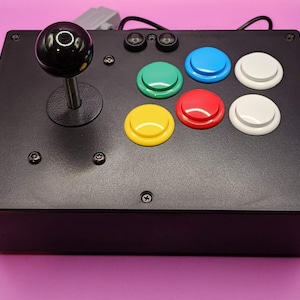 May include: Black arcade-style controller with a joystick and six colorful buttons: green, blue, red, yellow, and white. The joystick has a black ball top. The controller is on a pink surface.