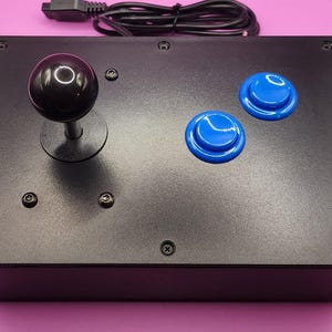 May include: A black arcade joystick with a black ball top and two blue buttons. The joystick is connected to a black cable.