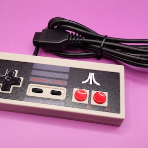 May include: A retro-style video game controller with a cream-colored body and a black control panel. The controller features a directional pad, two red buttons, and the Atari logo. A black cable extends from the top.