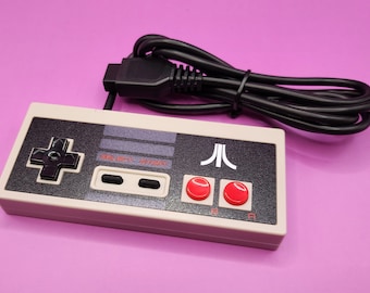 May include: A retro-style video game controller with a cream-colored body and a black control panel. The controller features a directional pad, two red buttons, and the Atari logo. A black cable extends from the top.