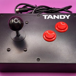 May include: A black TANDY joystick with two red buttons and a black knob. The joystick is connected to a black cable with a connector on the end.