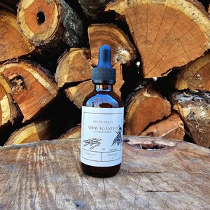 May include: A brown glass bottle with a black dropper and a white label. The label reads "River Roots Ashwagandha with Brandy & Honey" with a bee illustration. The bottle is on a wooden surface with a background of stacked firewood.