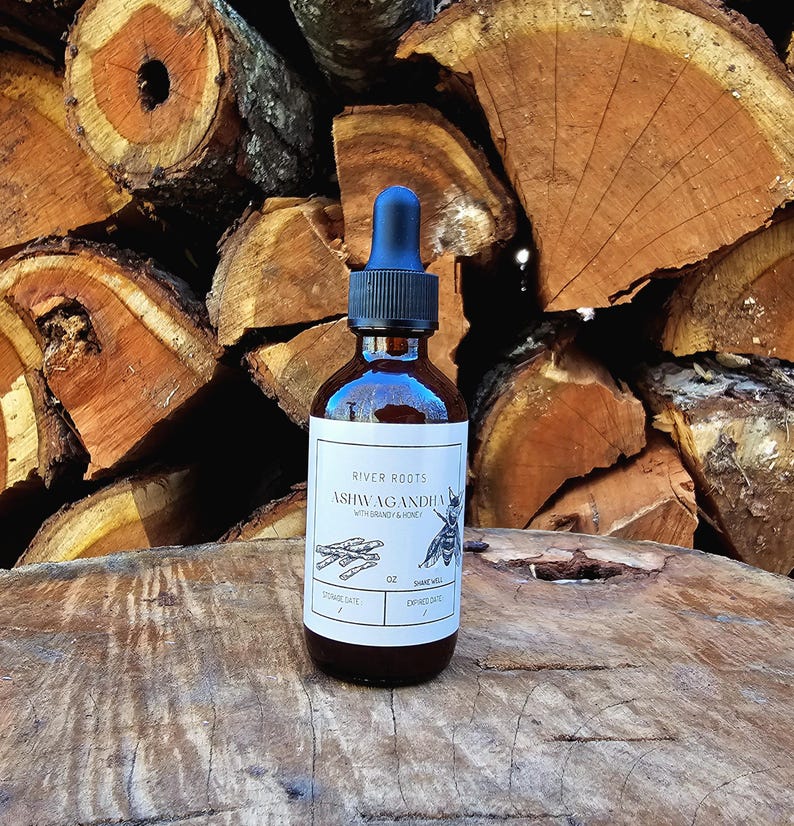 May include: A brown glass bottle with a black dropper and a white label. The label reads "River Roots Ashwagandha with Brandy & Honey" with a bee illustration. The bottle is on a wooden surface with a background of stacked firewood.