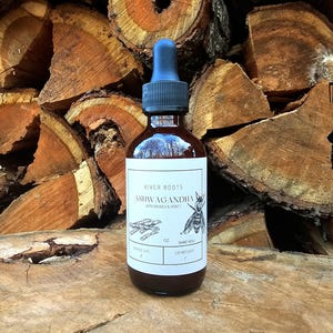 May include: A brown glass bottle with a blue dropper top. The label reads "River Roots Ashwagandha with Brandy & Honey." The label also features an illustration of a bee and plant roots. The bottle is sitting in front of a stack of wood.